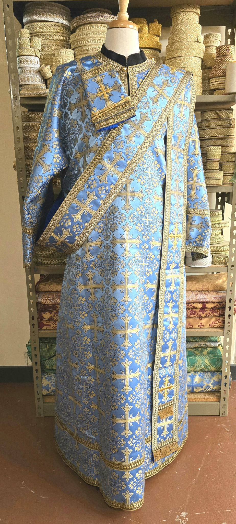Blue liturgical vestment with gold embroidery on a mannequin against a wooden shelf background.